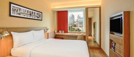 A hotel room with a large bed, a TV, a desk with a chair, a window with a city view, and a framed picture on the wall.