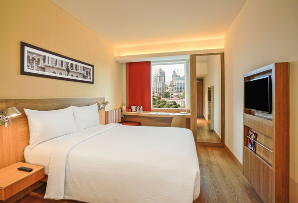 Une chambre d’hôtel avec un grand lit, une télévision, un bureau avec une chaise, une fenêtre donnant sur la ville et un tableau encadré au mur.