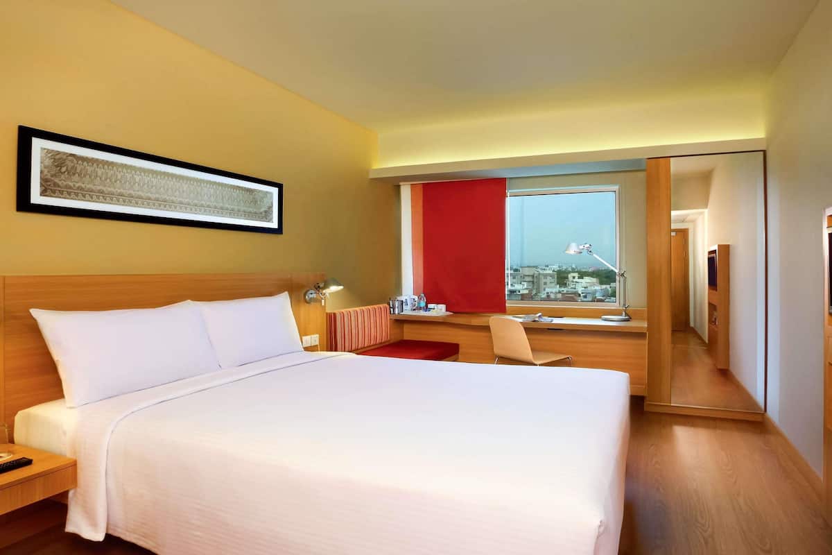 A hotel room with a large bed, a desk with a chair, a window with a red curtain, and a city view.