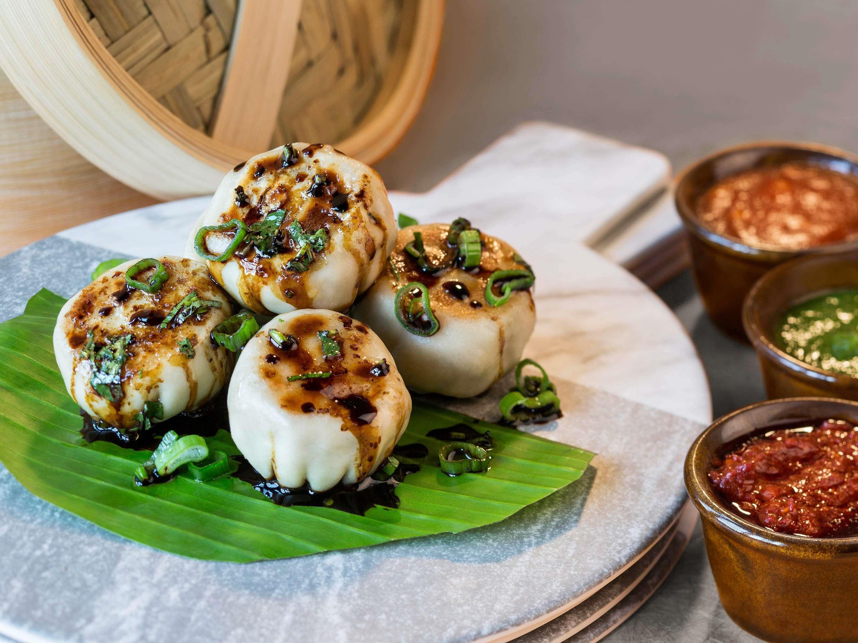 A plate of steamed buns garnished with green onions and sauce, accompanied by two small bowls of dipping sauce.
