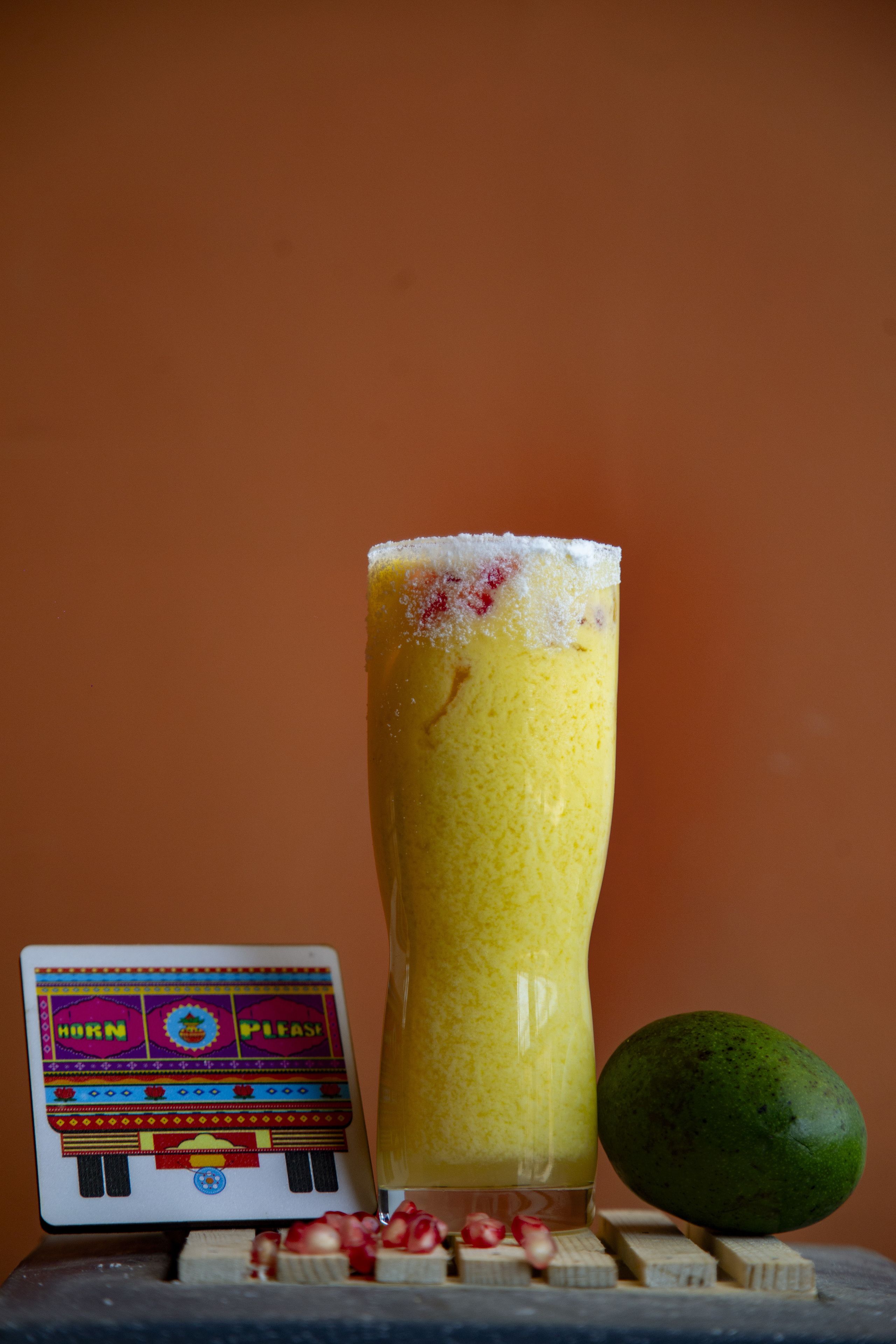 A tall glass of yellow beverage with a frothy top, a green fruit, and a small card with colorful patterns.
