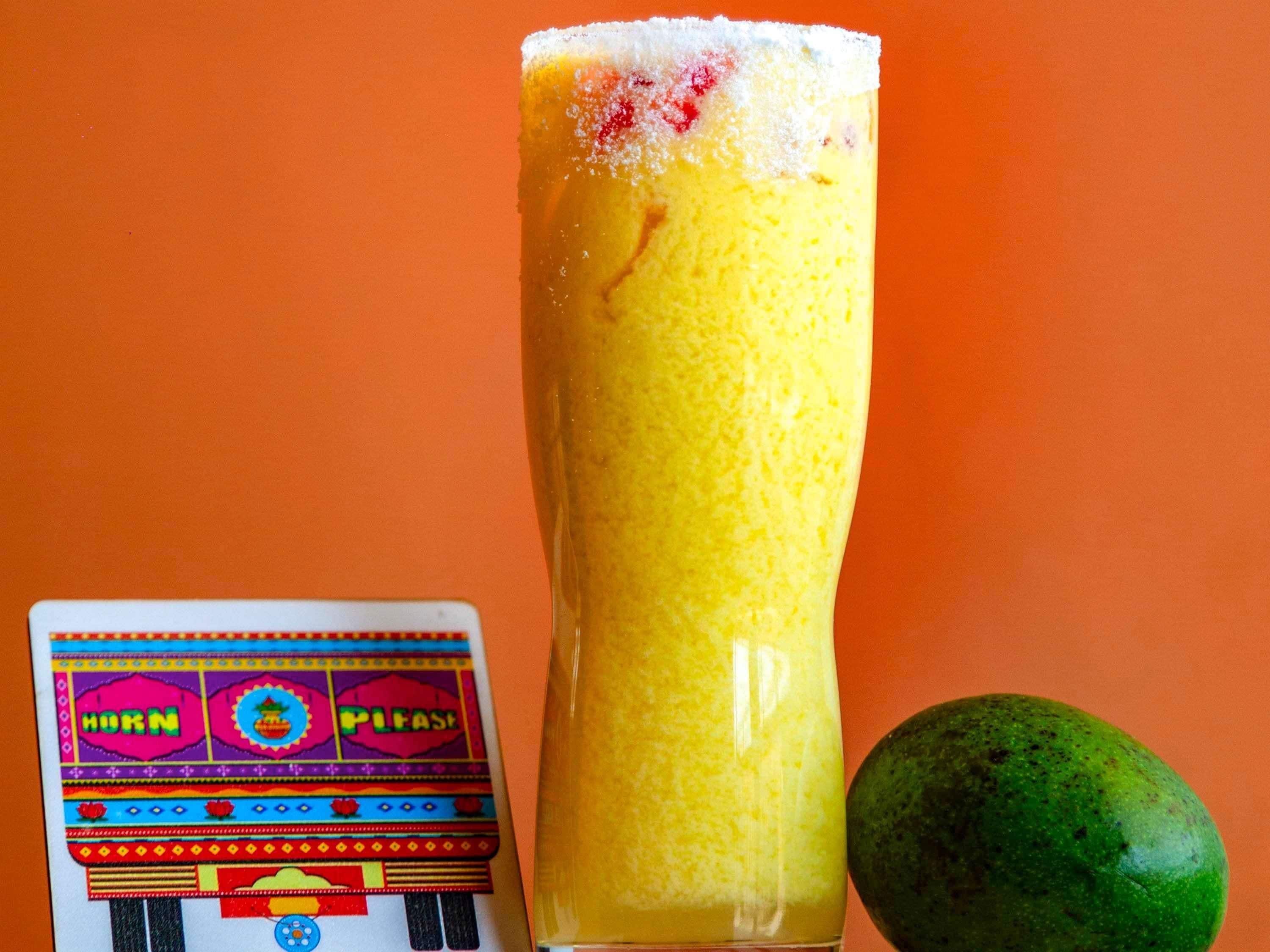 A tall glass of yellow beverage with a frothy top, a colorful card, and a green fruit beside it.