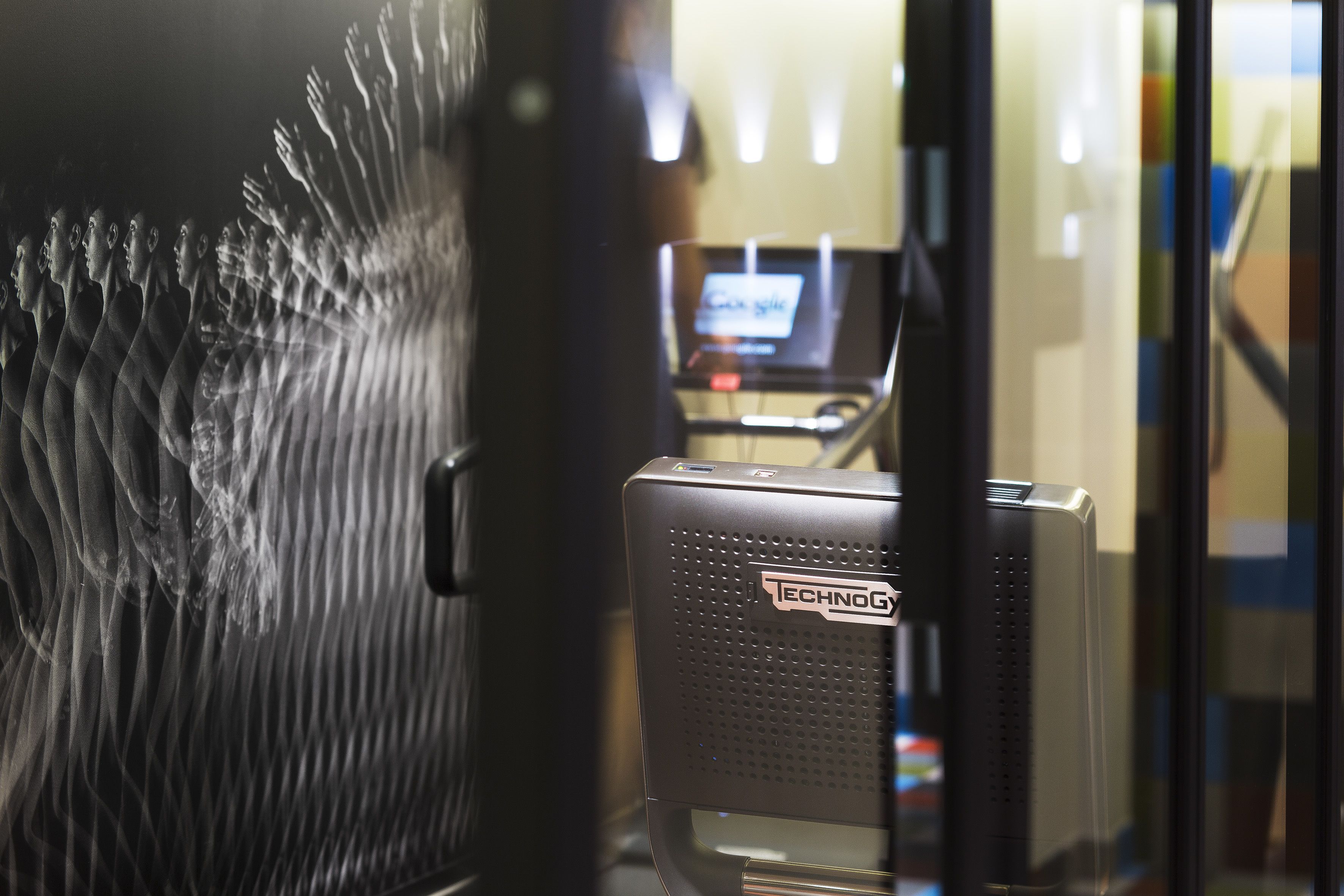 A view through a glass door with a reflection of a room containing a desk and a computer.