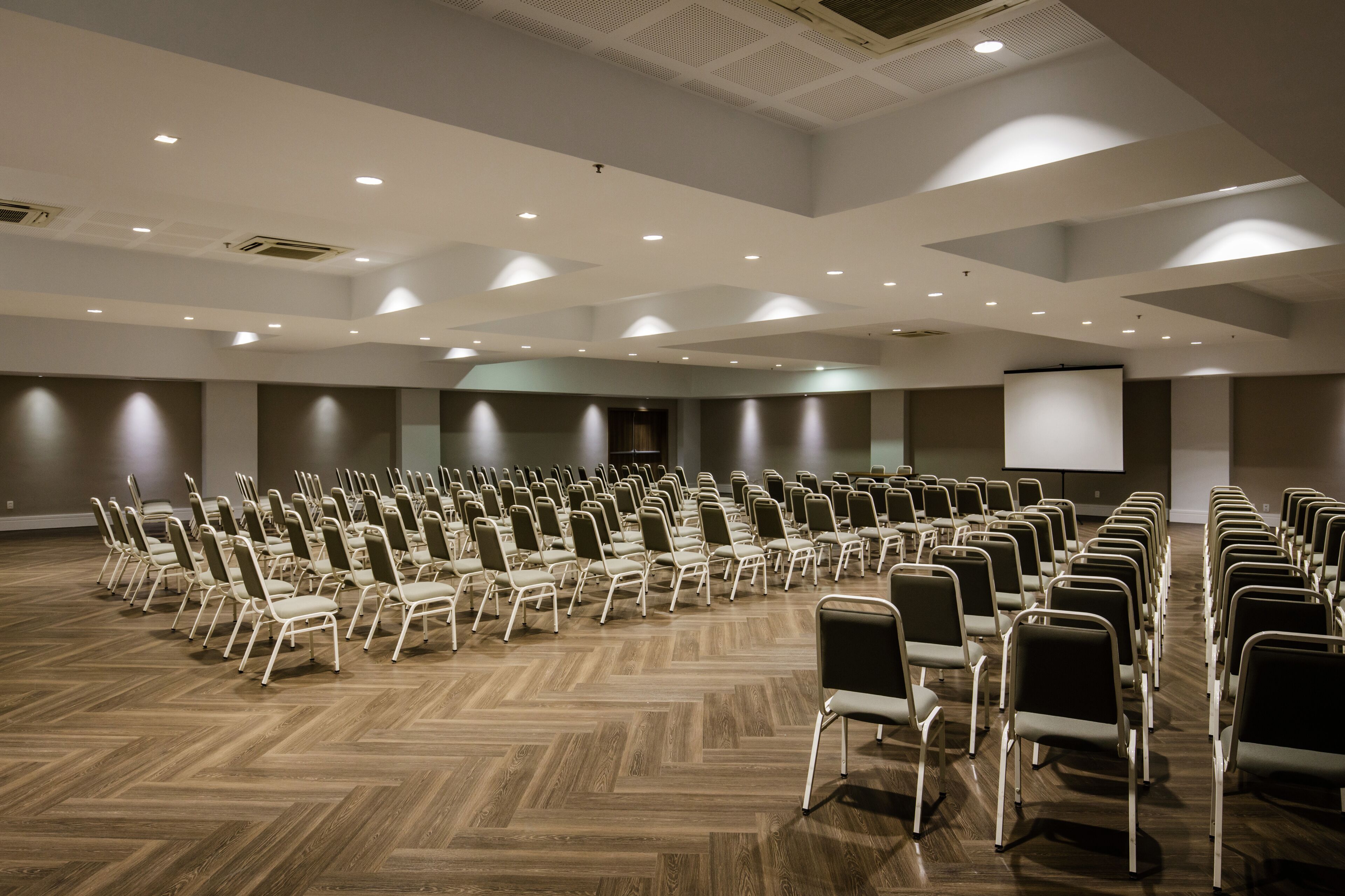 A conference room with rows of chairs, a projector screen, and a wooden floor.