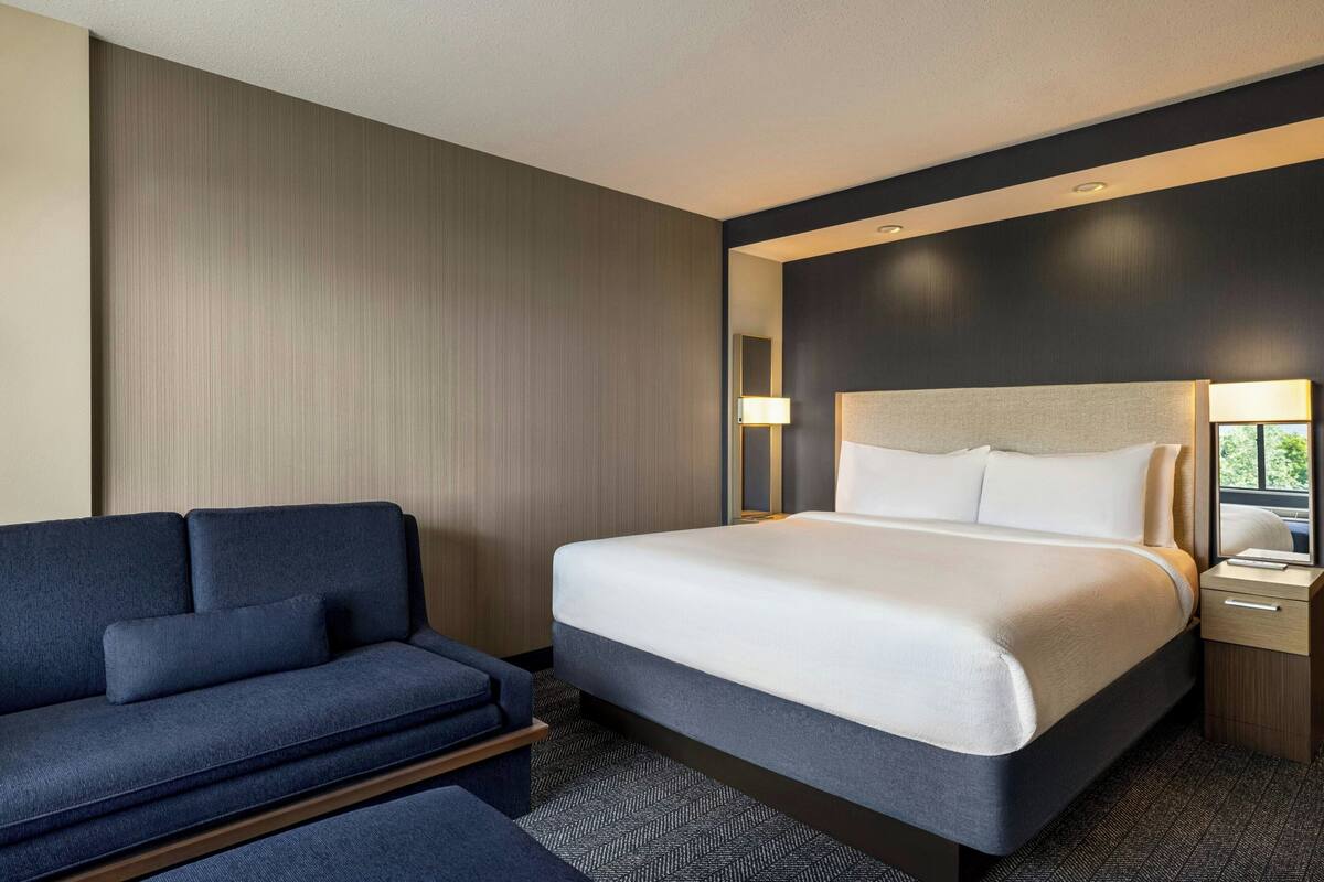 A modern hotel room with a large bed, a sofa, a bedside table, and a lamp.