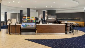 A modern hotel lobby with a central reception desk, a beverage station, and seating areas.