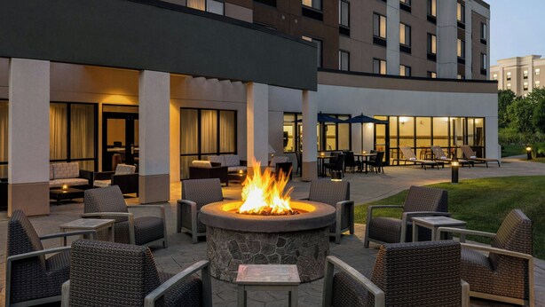 An outdoor seating area with a stone fire pit surrounded by wicker chairs, a covered patio with tables and chairs, and a building with multiple windows and balconies in the background.