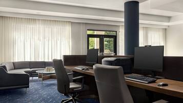 A modern hotel room with a large desk, two monitors, a keyboard, and a mouse. There is a sofa and a coffee table.