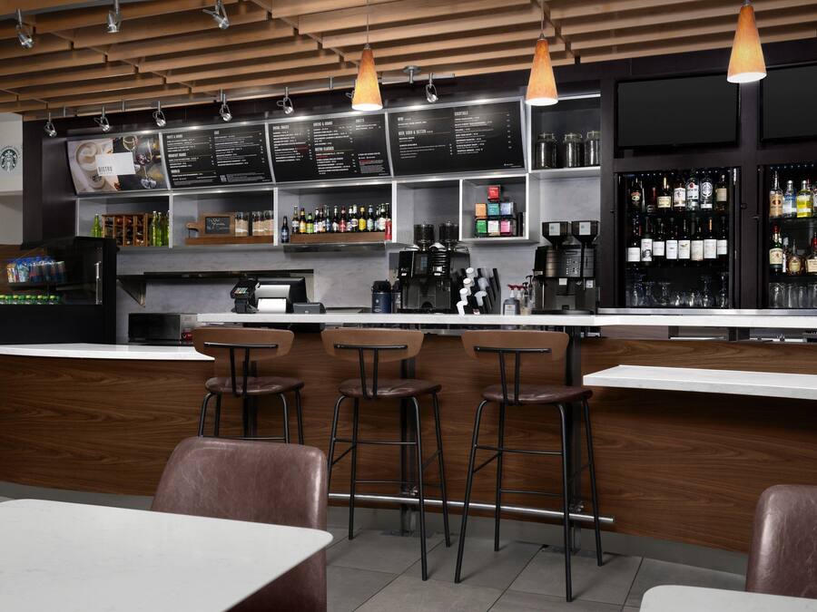 A modern bar area with wooden ceiling, bar stools, a coffee machine, and shelves stocked with bottles.