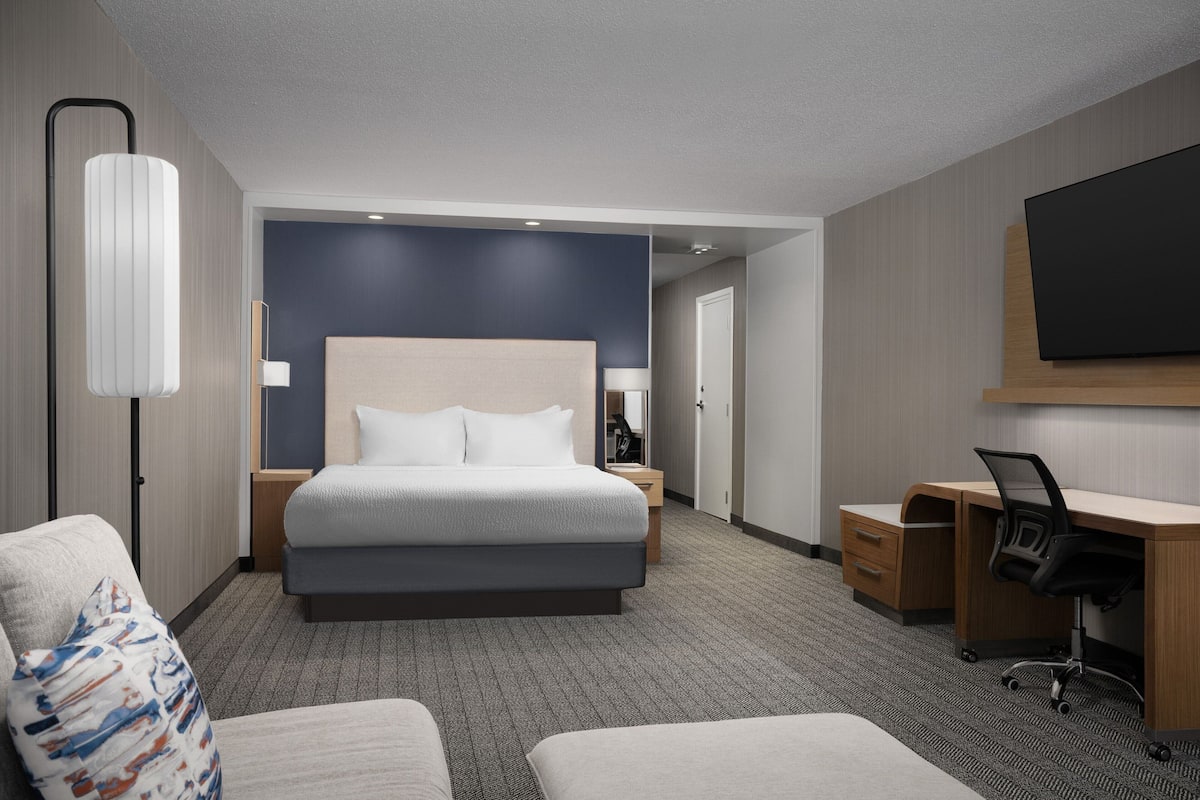A hotel room with a large bed, a desk, a chair, a TV, and a lamp.