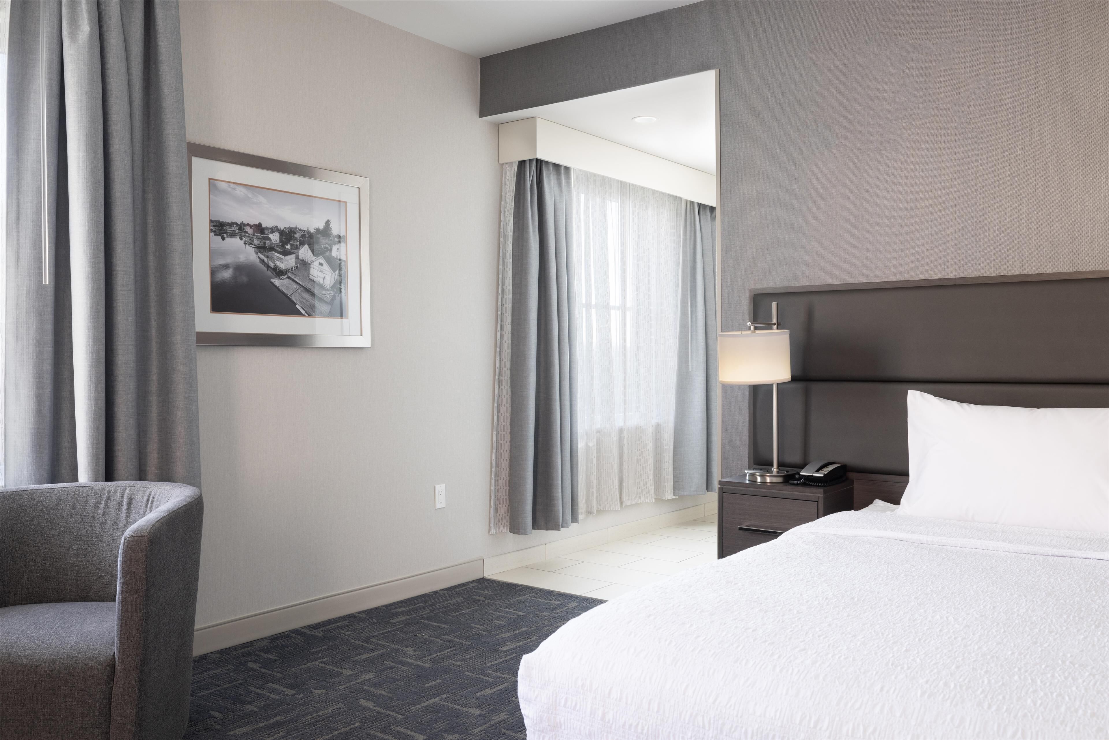 A hotel room with a large bed, a chair, a nightstand with a lamp, and a framed picture on the wall.