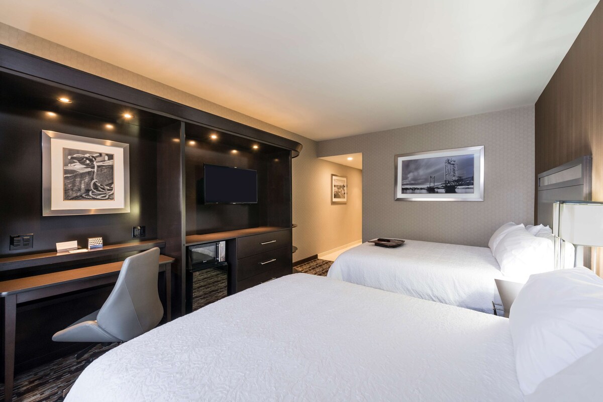 A hotel room with two beds, a desk, a TV, and artwork on the walls.