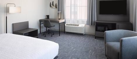 A hotel room with a bed, a desk, a chair, a TV, and a heater.