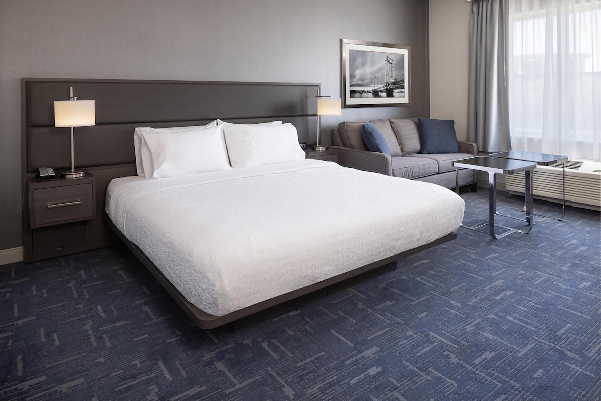 Ein Hotelzimmer mit einem großen Bett, einem Nachttisch mit Lampe, einem Sofa, einem kleinen Tisch und einem Fenster mit Vorhängen.