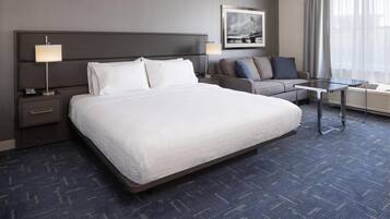 Una habitación de hotel con una cama grande, una mesita de noche con lámpara, un sofá, una pequeña mesa y una ventana con cortinas.