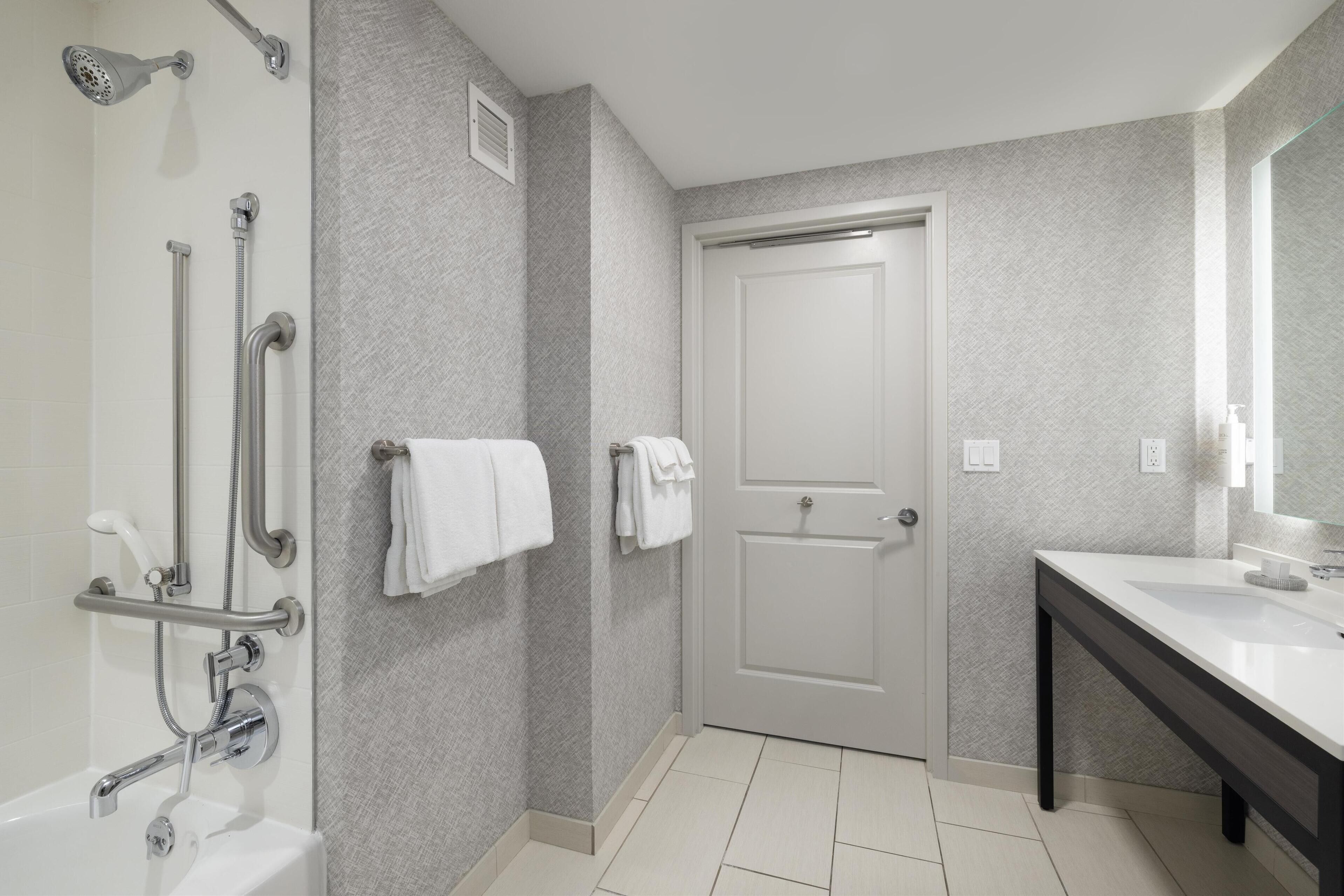 Un baño moderno con ducha, barras de apoyo, una puerta blanca y un lavamanos con espejo.