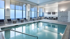 An indoor swimming pool with clear glass walls, lounge chairs, and a view of the ocean.