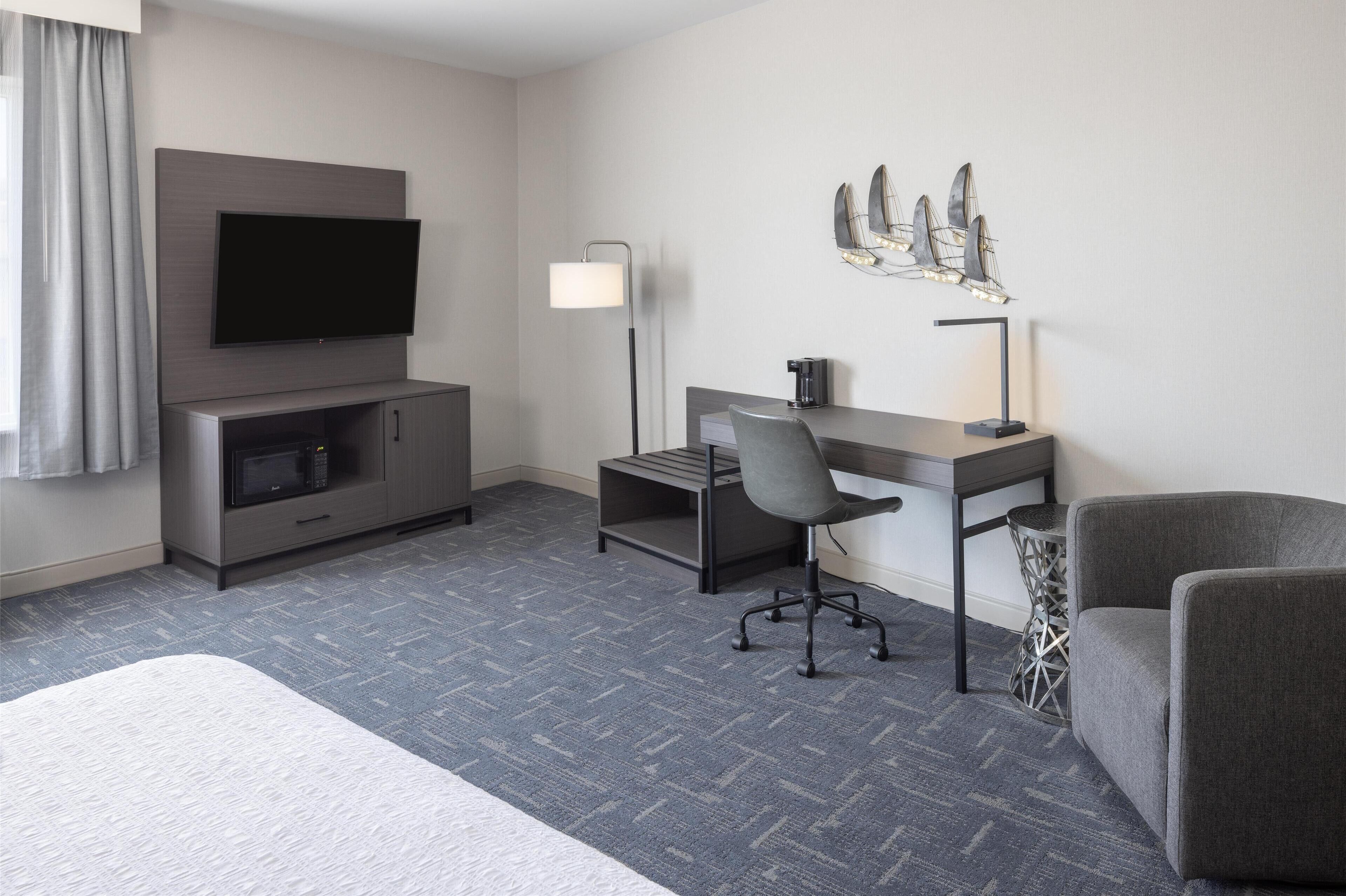 A modern hotel room with a flat-screen TV, a desk with a chair, a lamp, and a decorative wall piece.