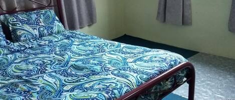 Una cama con una colcha estampada, estructura metálica y cortinas en una ventana.
