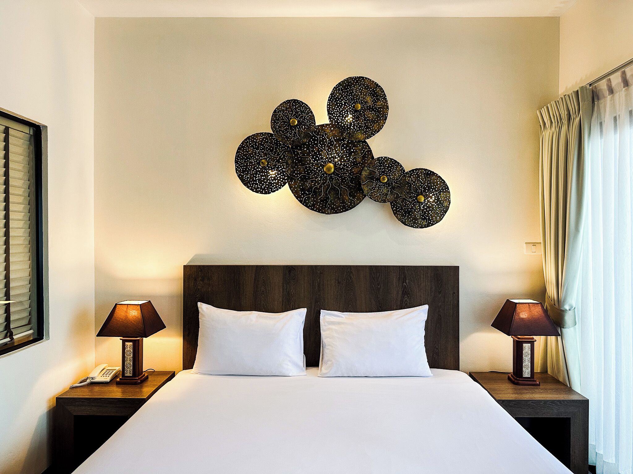 A hotel room with a large bed, two bedside tables with lamps, a decorative wall piece, and a window with curtains.