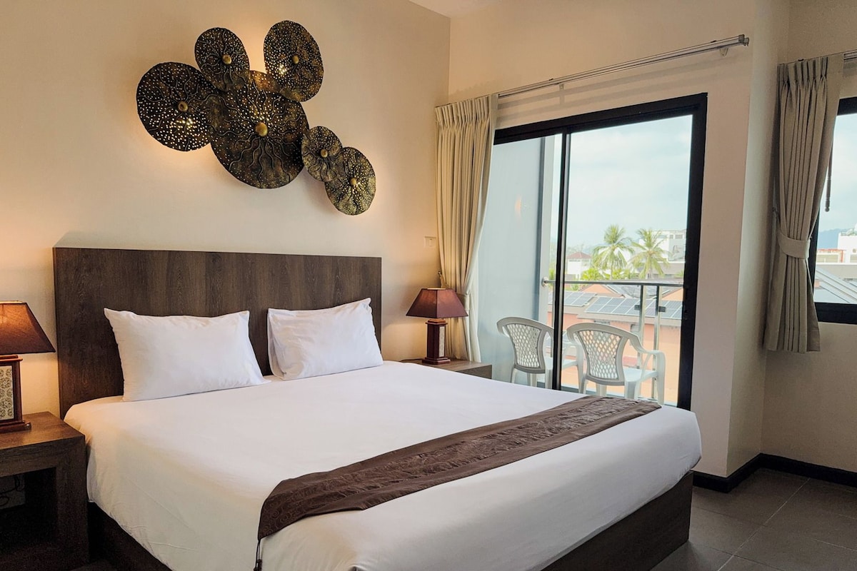 A hotel room with a large bed, a nightstand, a lamp, a decorative wall piece, and a view of the outside through a sliding door.