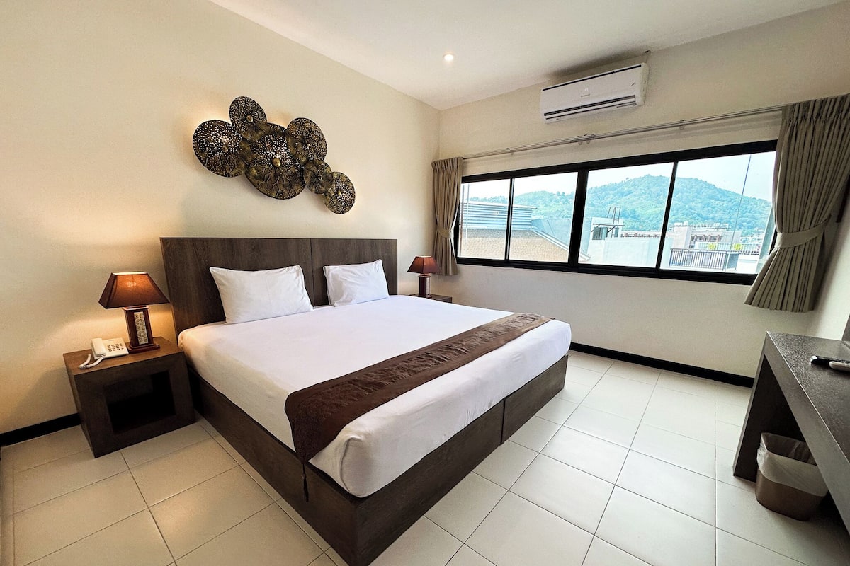 A hotel room with a large bed, a nightstand with a lamp, a desk with a chair, and a window with a view of the city.