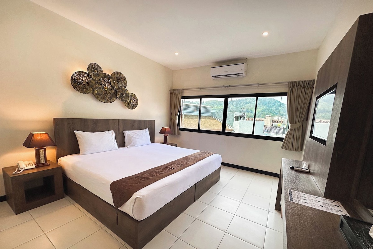 A hotel room with a large bed, a bedside table with a lamp, an air conditioning unit, a window with a view, and a curtain.
