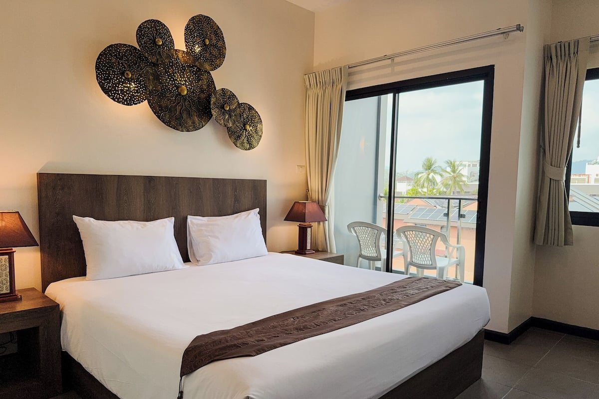 A hotel room with a large bed, a nightstand, a lamp, a wall art piece, a balcony with chairs, and a view of the cityscape.
