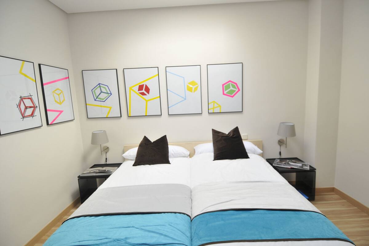 A hotel room with a large bed, two bedside tables, a nightstand, and five framed geometric art pieces on the wall.