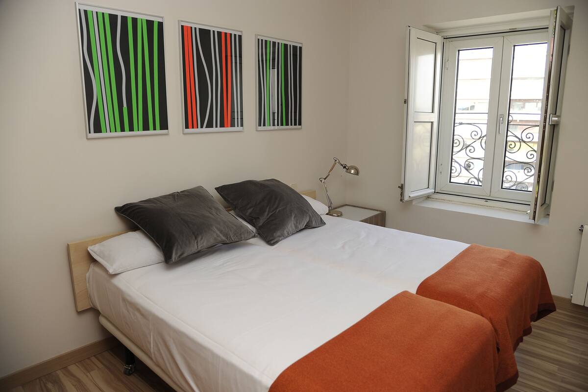 A bedroom with a bed, two pillows, a bedside table with a lamp, and a window with a balcony view.