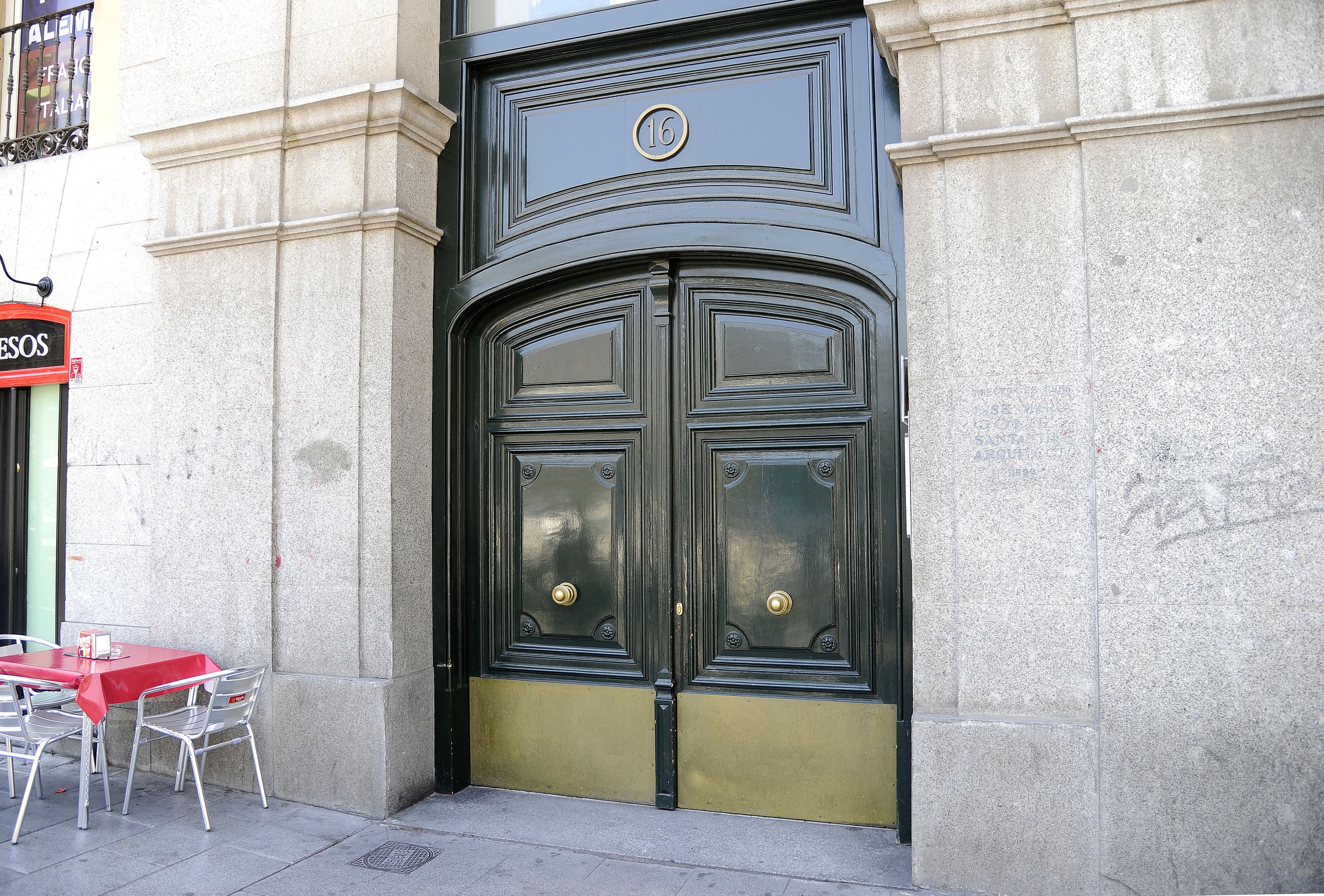 Une porte à deux battants, dotée d’une poignée en laiton et d’un emblème circulaire au-dessus, est intégrée à un bâtiment en pierre. Devant, on aperçoit une table et des chaises rouges.
