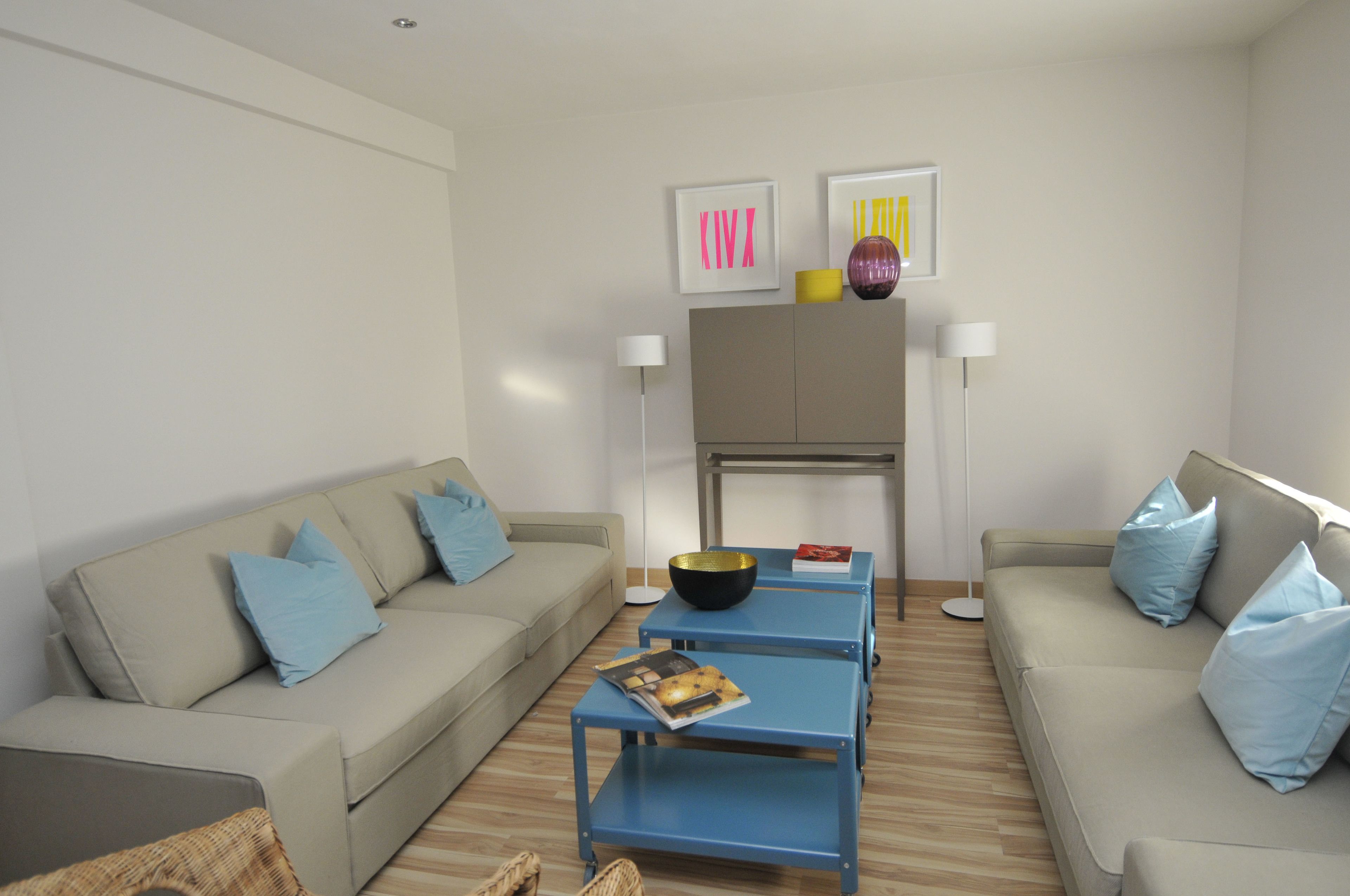 A modern living room with two sofas, a coffee table, and a small dining table. There are two wall-mounted lamps and framed artwork above the dining table.