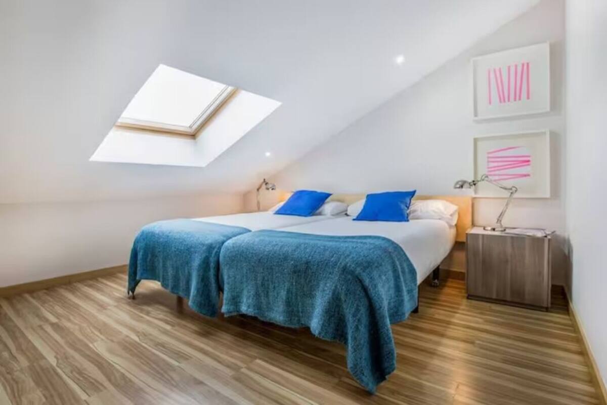 A bedroom with a skylight, a bed with blue and white bedding, a nightstand, and a wall with abstract art.