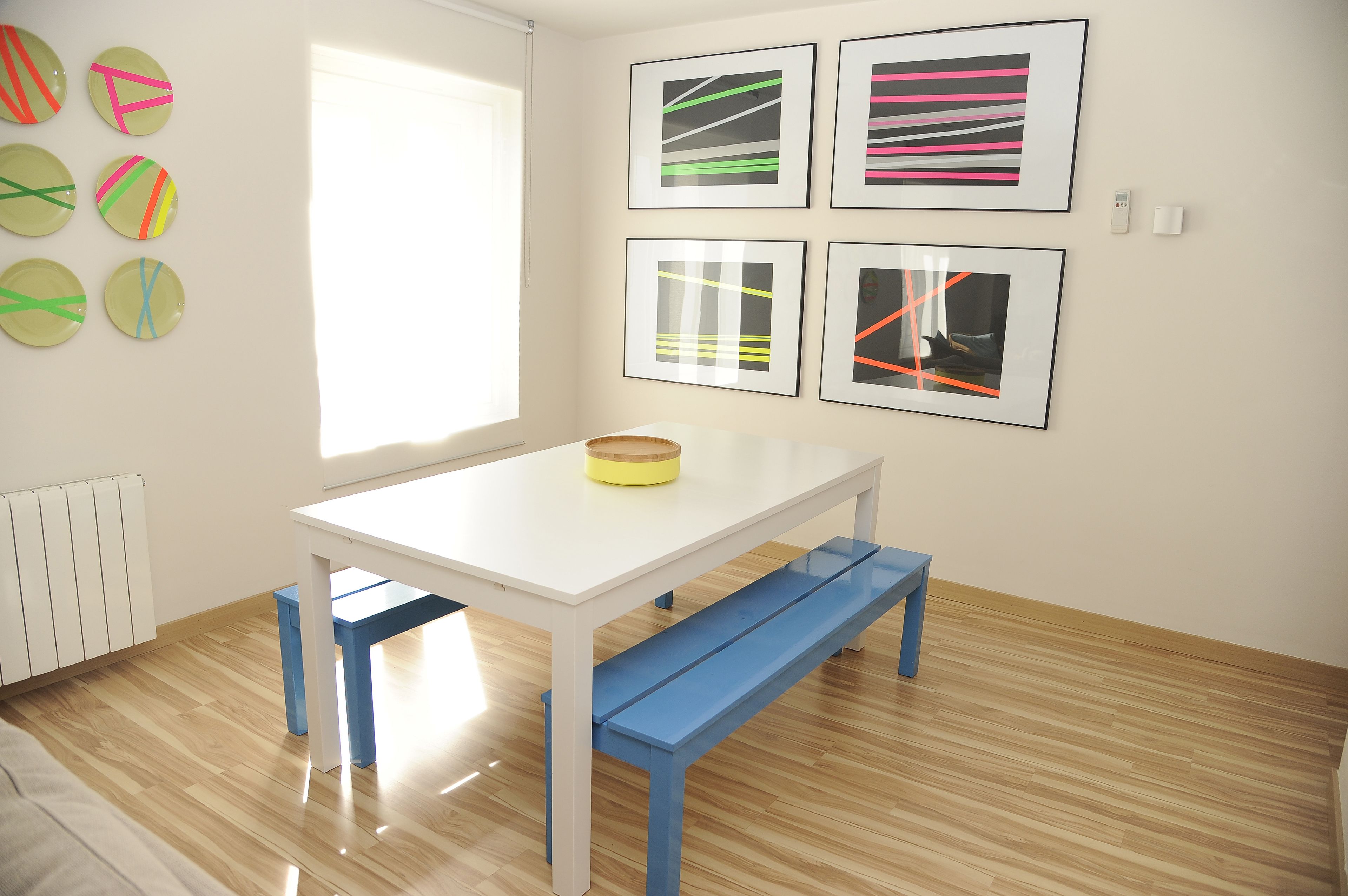 Una stanza con un tavolo bianco e panche blu, un termosifone e sei opere d'arte incorniciate appese al muro.
