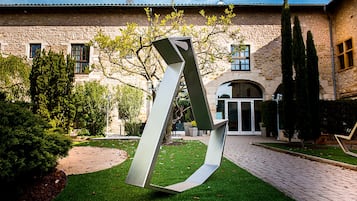 A courtyard with a modern metal sculpture, a stone building with arched windows, and well-maintained greenery.