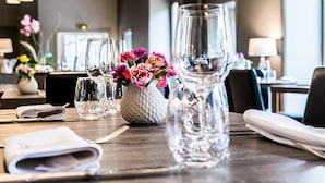 Ein Esstisch ist gedeckt mit klarem Glas, einer weißen, strukturierten Vase mit rosa Blumen und ordentlich gefalteten Servietten.