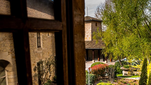 Blick durch ein Fenster auf einen Innenhof mit einem Steinhaus, einem gepflegten Garten und einer Schaukel.