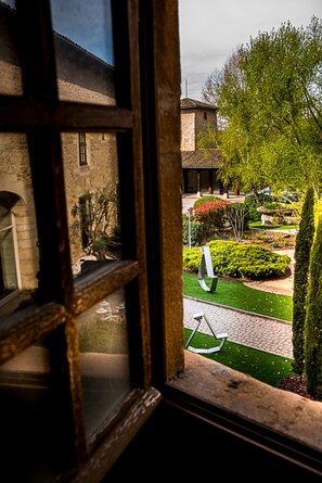 Une vue à travers une fenêtre donnant sur une cour intérieure, avec un bâtiment en pierre, un jardin bien entretenu et une balançoire.