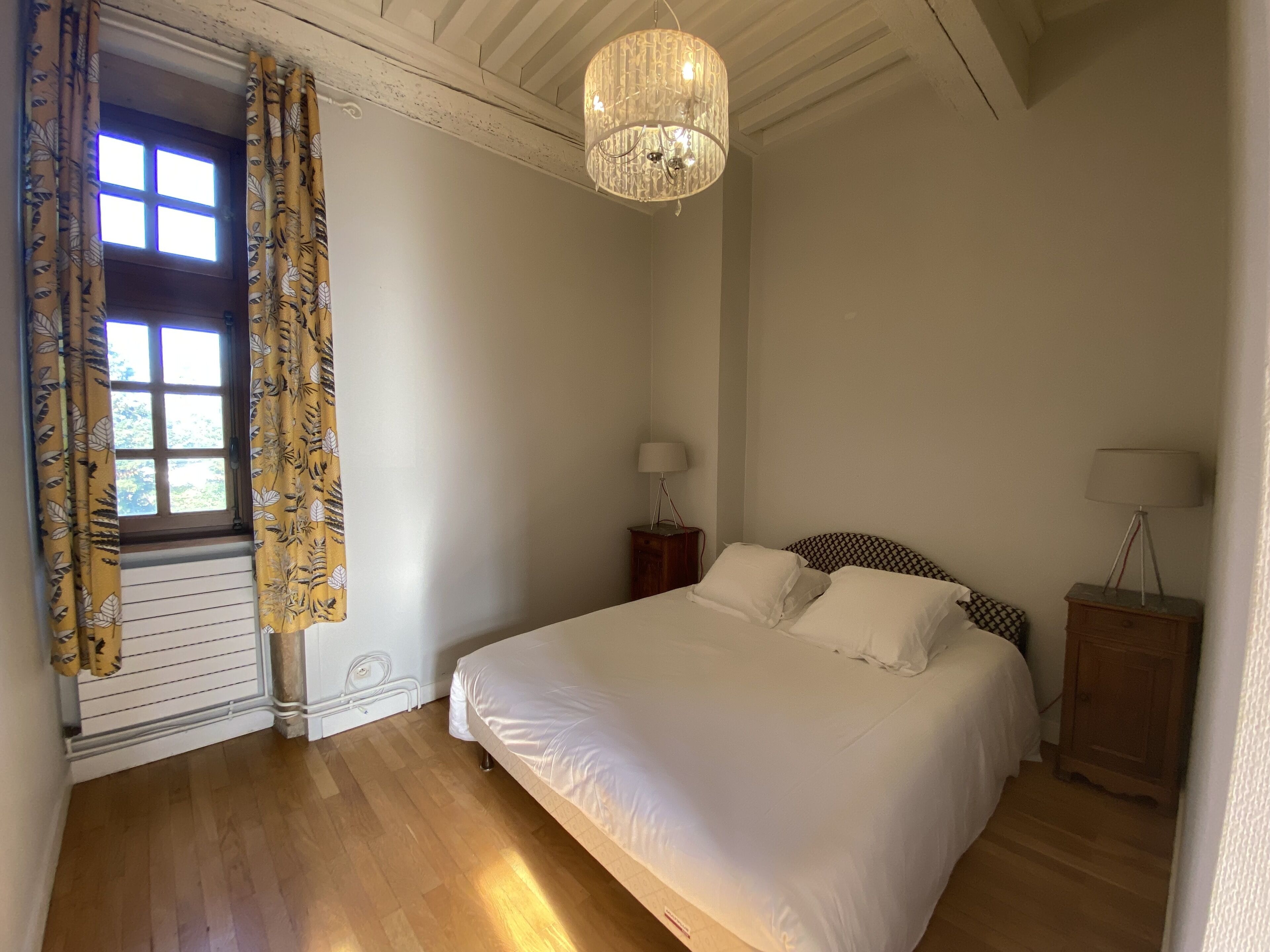 Une chambre à coucher avec un grand lit, un lustre, une fenêtre avec des rideaux et une table de chevet avec une lampe.