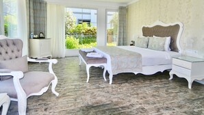 A bedroom with a large bed, a chaise lounge, a nightstand, and a view of greenery outside.