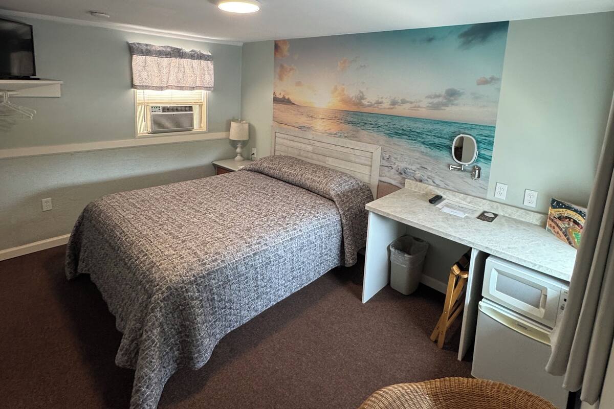 A bedroom with a bed, a desk, a chair, a TV, and a beach mural.