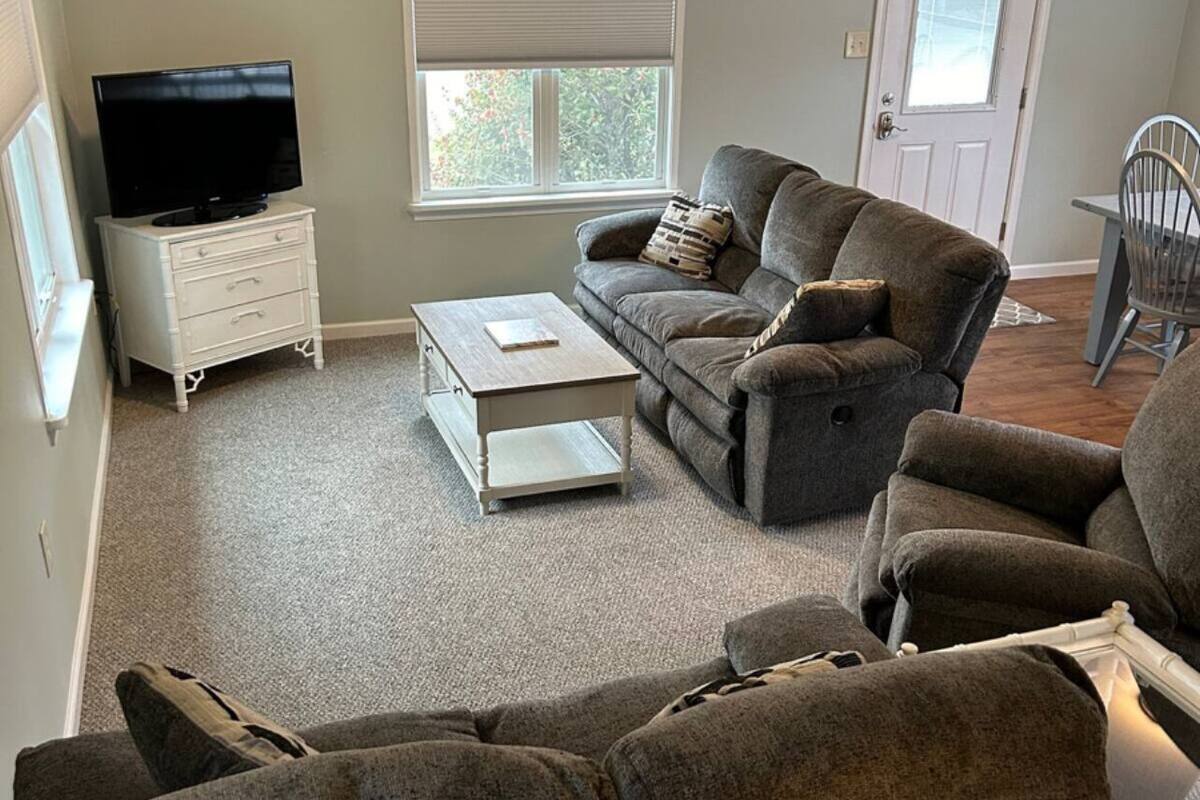 A living room with a grey sofa, a coffee table, a TV on a white cabinet, a ceiling fan, and a window with blinds.