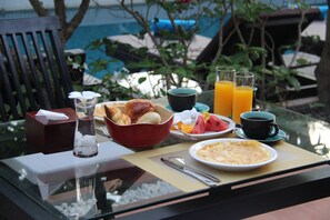 A breakfast table with various food items, a glass of orange juice, and a view of the pool.