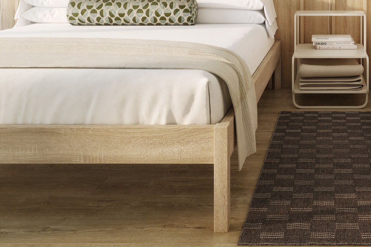 Quarto com cabeceira de madeira, cama com roupa de cama branca, almofada estampada, mesa de cabeceira com abajur e uma pequena estante.
