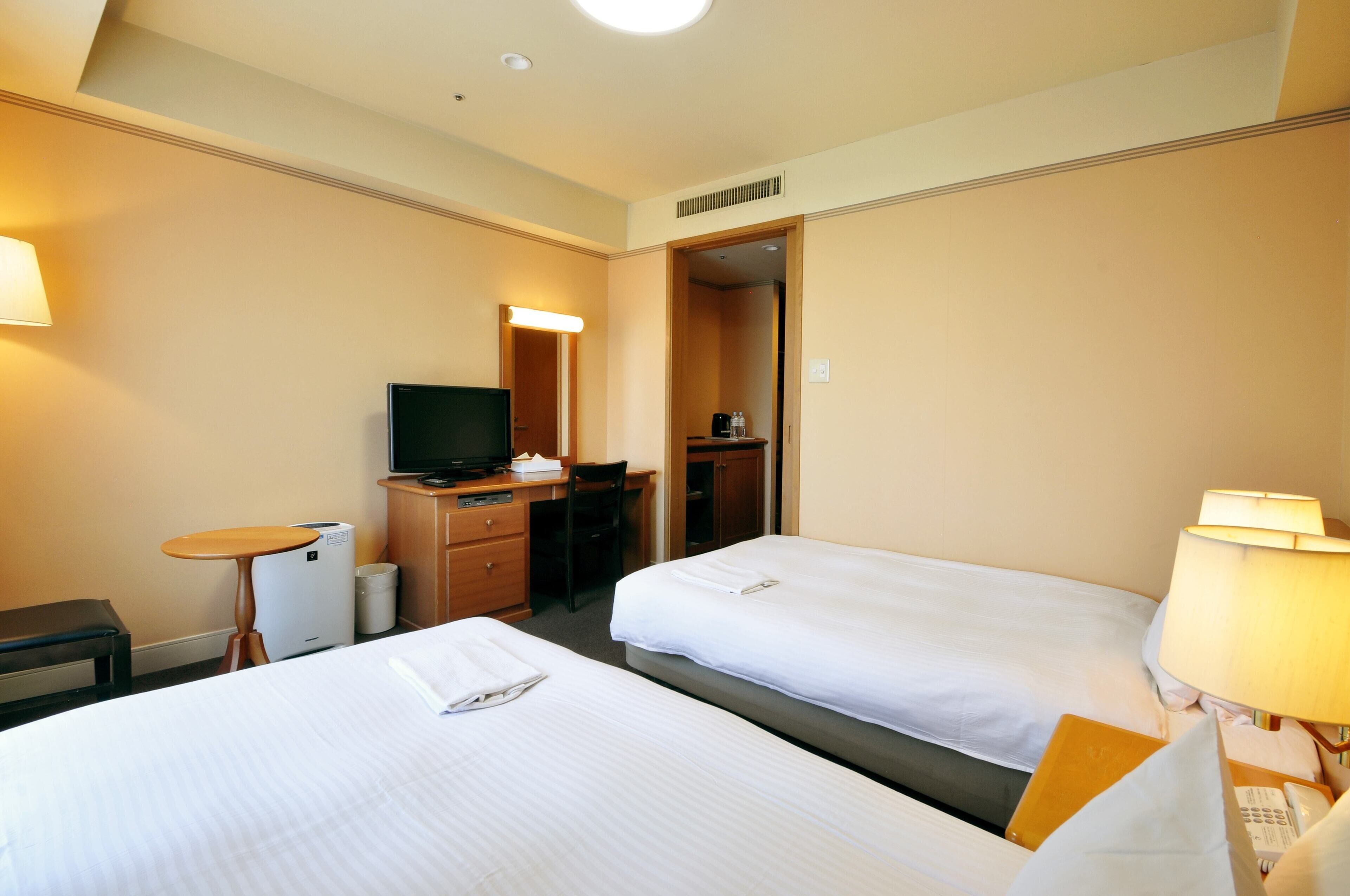 A hotel room with two beds, a desk with a computer, a television, and a small table with a lamp.