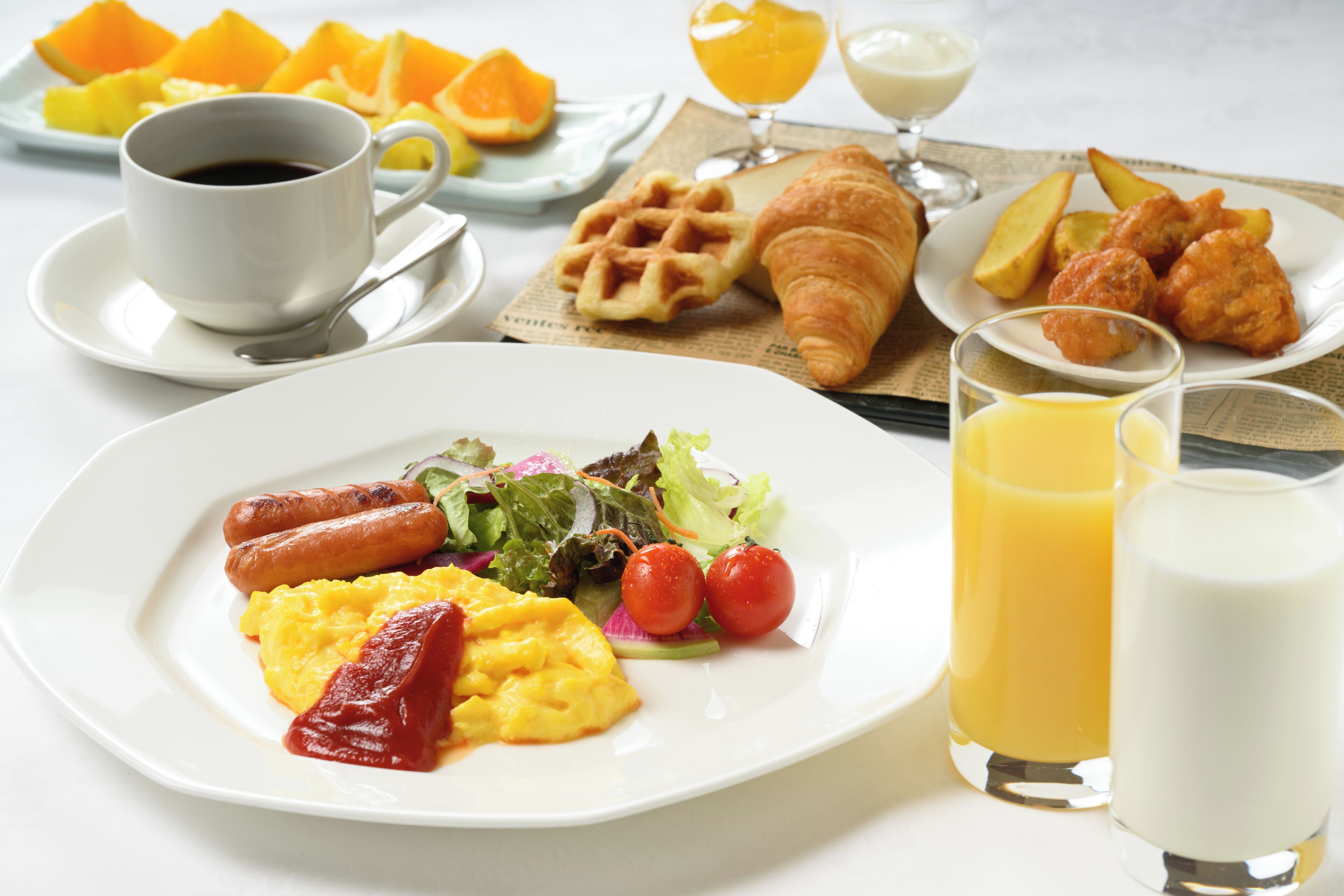 A breakfast plate with sausages, scrambled eggs, and a salad, accompanied by a cup of coffee, a waffle, croissants, fried chicken, and orange juice.