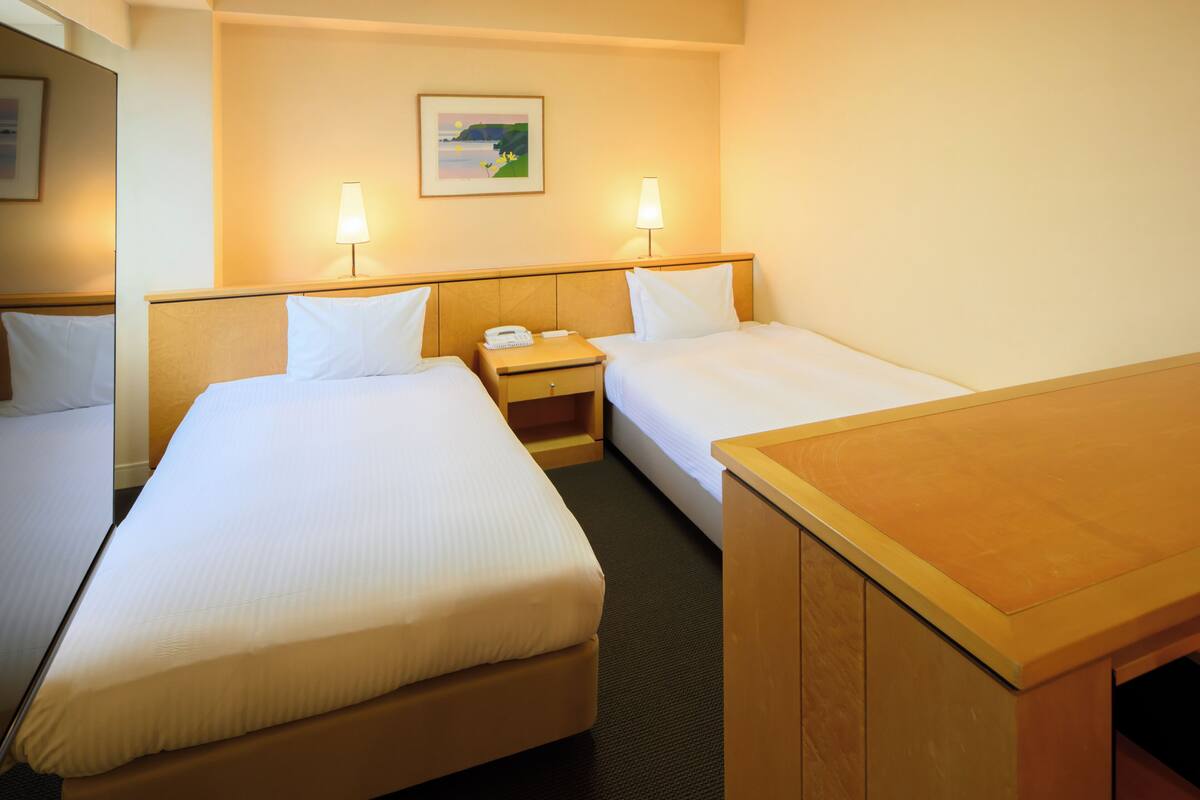 Habitación de hotel con dos camas, un escritorio de madera, una mesita de noche con lámpara y un cuadro enmarcado en la pared.