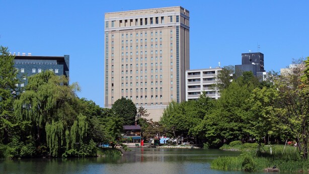 A tall hotel with a modern design, surrounded by lush greenery and a body of water. - Hotel Lifort Sapporo (Sapporo)
