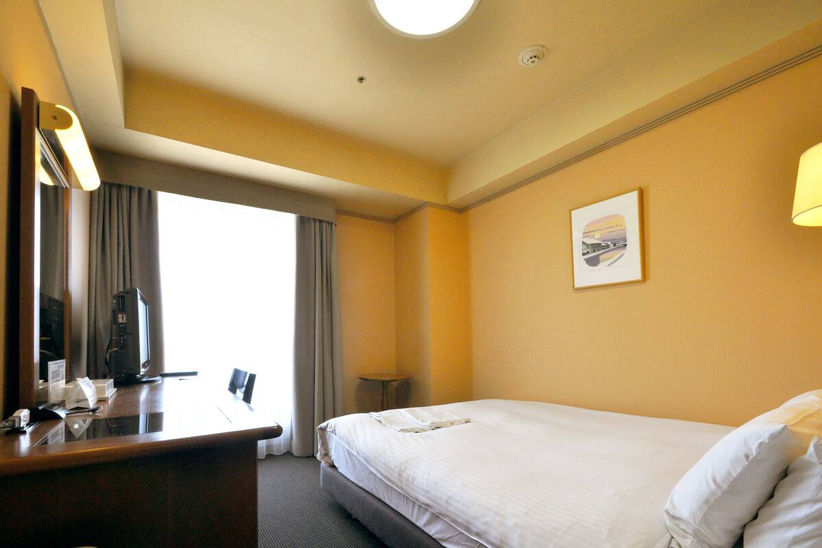 Habitación de hotel con cama, escritorio, computadora, lámpara y ventana con cortinas.