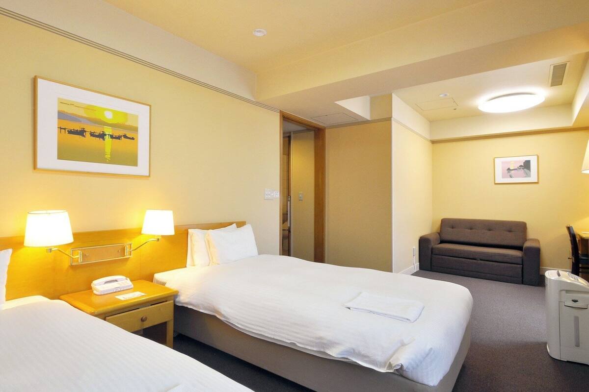A hotel room with two beds, a chair, a desk with a lamp, and a painting on the wall.